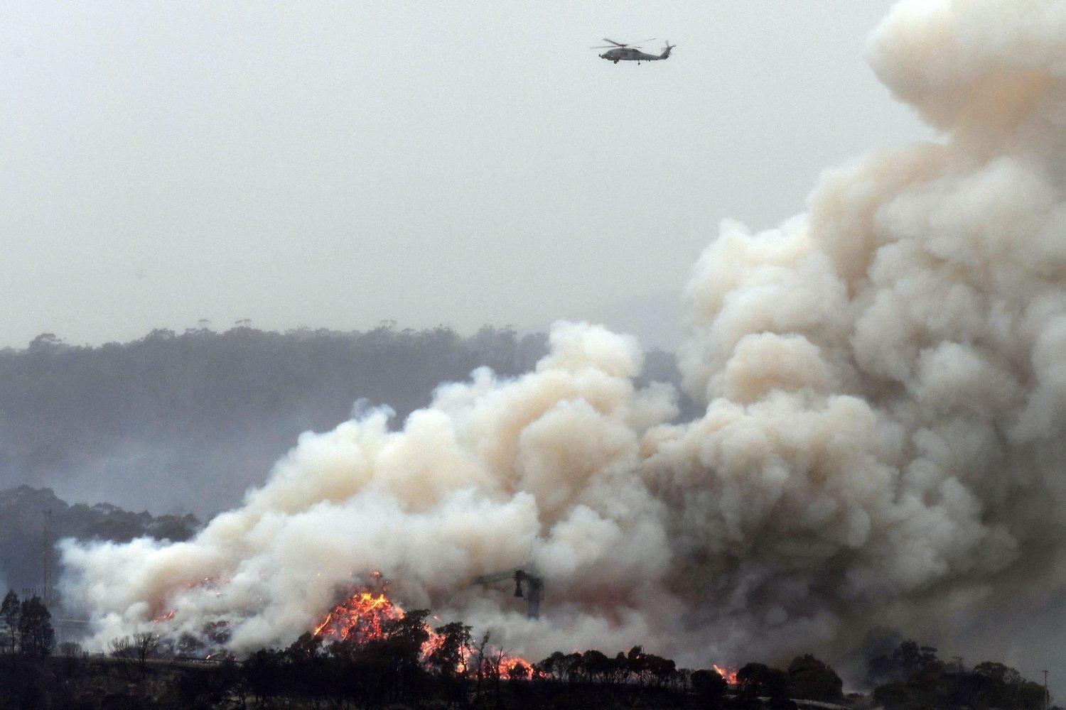 A military helicopter flies above a burning woodchip mill in Eden, in Australia's New South Wales state on January 6, 2020. - January 5 brought milder conditions, including some rainfall in New South Wales and neighbouring Victoria state, but some communities were still under threat from out-of-control blazes, particularly in and around the town of Eden in New South Wales near the Victorian border. (Photo by SAEED KHAN / AFP)